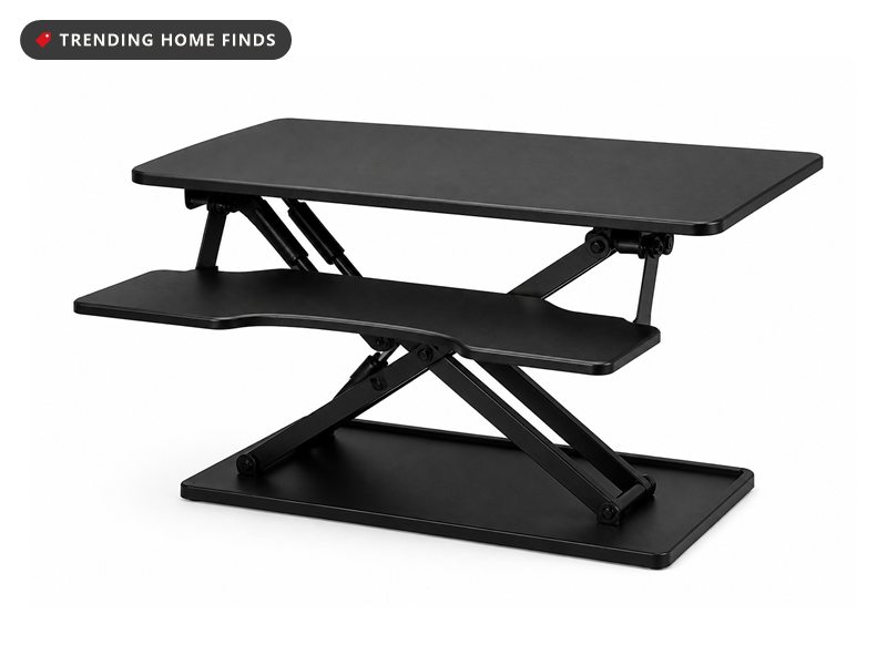 A black sit stand riser raised on a scissor frame with a wide top and a lower keyboard tray.