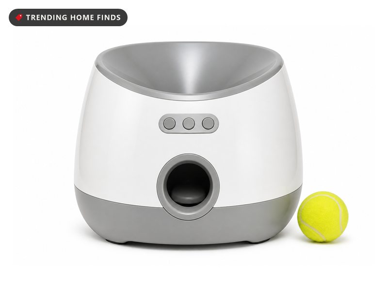 A white and gray automatic ball launcher with a funnel top and a front launch opening beside a tennis ball.