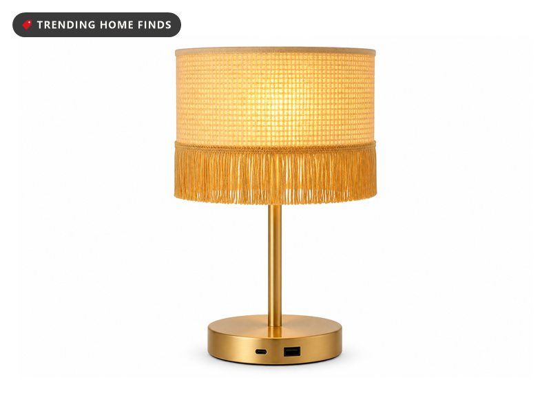 A fringed table lamp with a gold base, a woven cream shade and a long gold tassel fringe, switched on.