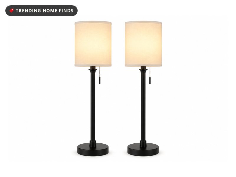Two matching tall slim black candlestick lamps with white linen drum shades, lit.