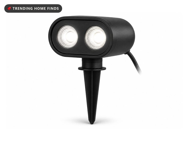 A compact black outdoor projector with two glowing round lenses on a short ground stake.