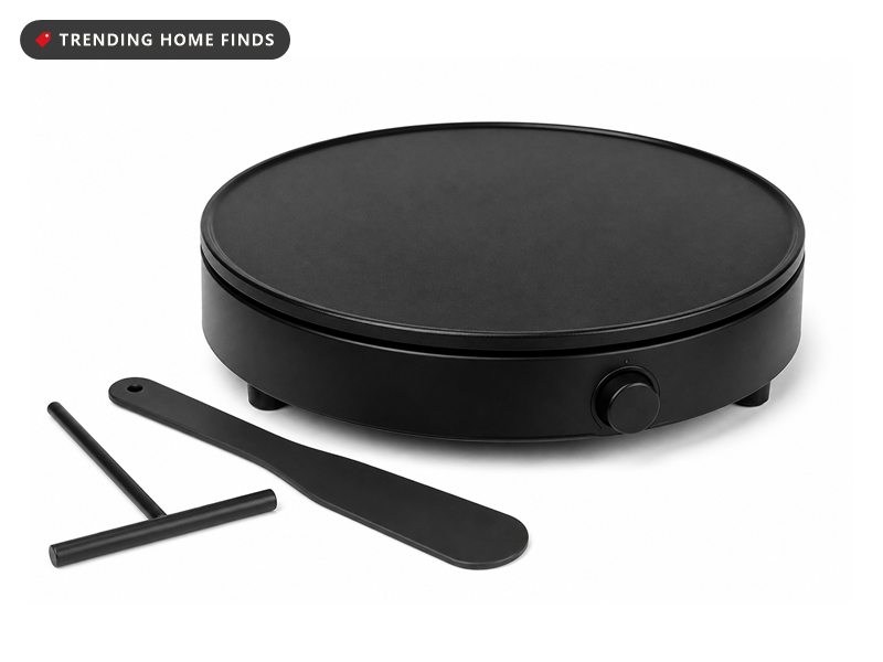 A round electric crepe maker with a flat black nonstick plate on a low base, a thin spreader and a spatula beside it.
