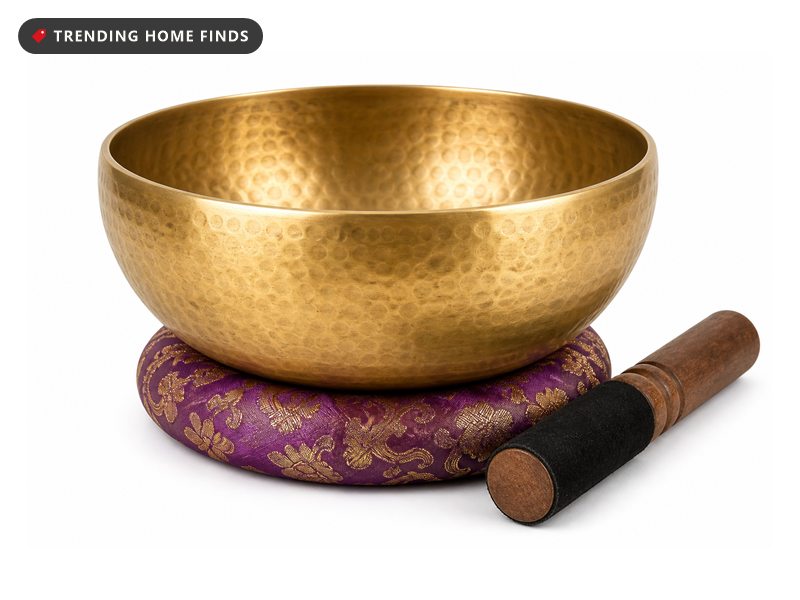 A hammered brass singing bowl on a round silk cushion with a wooden striker lying beside it.