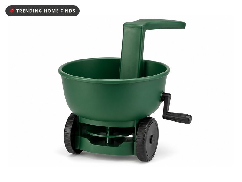 A green handheld crank spreader with a round open hopper, a side crank and a curved arm rest.