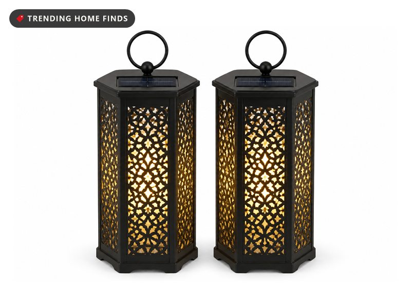 Two tall six sided black iron solar lanterns with pierced panels and loop handles, glowing warm white.