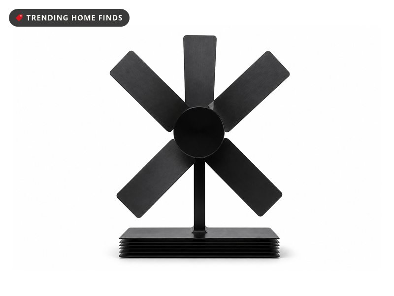 A black four blade stove fan on a finned base beside a small magnetic thermometer.