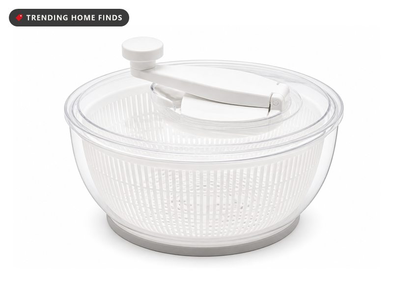 A white salad spinner with a clear lid, an extended folding crank handle and a gray non slip base ring.