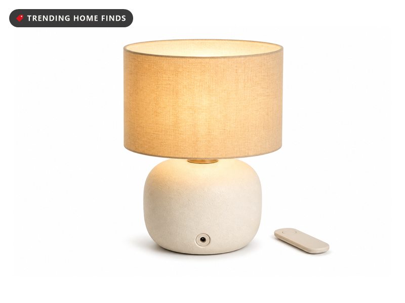A small cordless lamp with a rounded cream ceramic base and a beige linen shade glowing warm, a slim remote beside it.