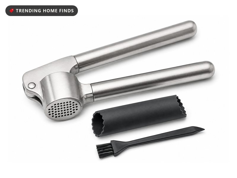 A heavy brushed stainless steel garlic press with long handles, a silicone tube peeler and a small cleaning brush beside it.